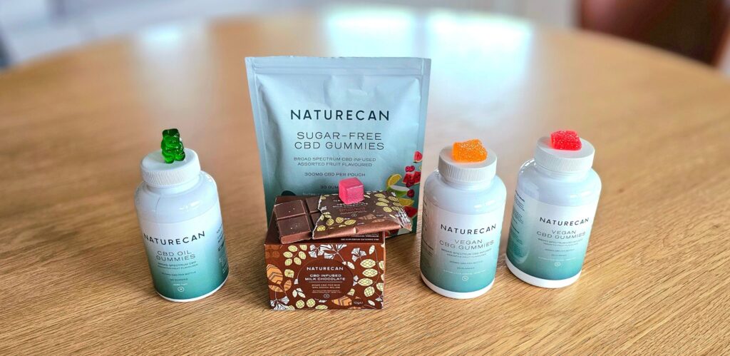 CBD godis från Naturecan står uppsradat på ett bord. Gummies står på burkarnas lock och i mitten lutar en chokladkaka.