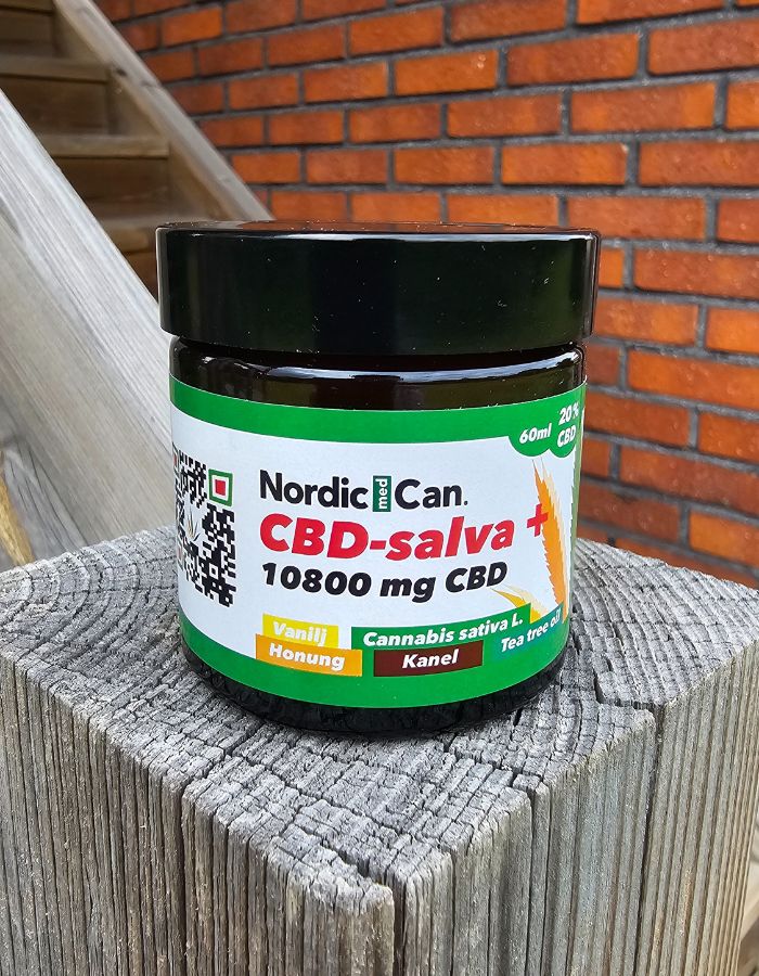 En burk med Nordic Med Can CBD salva + 10800 mg CBD. Burken har svart lock och en vit-grön etikett med gula och röda detaljer. Burken står placerad på en trästolpe med en tegelvägg i bakgrunden.