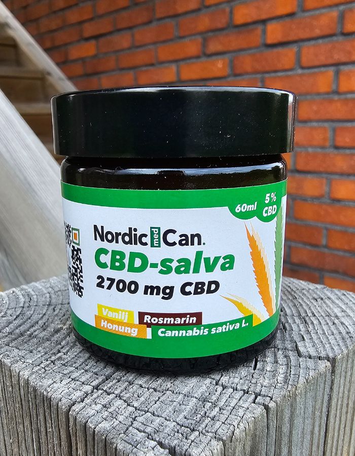En burk med Nordic Med Can CBD salva 2700 mg CBD. Burken har svart lock och en vit-grön etikett med gula och svarta detaljer. Burken står placerad på en trästolpe med en tegelvägg i bakgrunden.