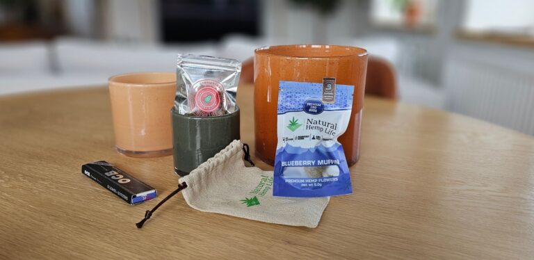 En hemtrevlig ställning med Natural Hemp Life's CBD-buds, Strawberry Diesel och Blueberry Muffin förpackningar, bredvid en hampapåse och rullpapper på ett träbord.