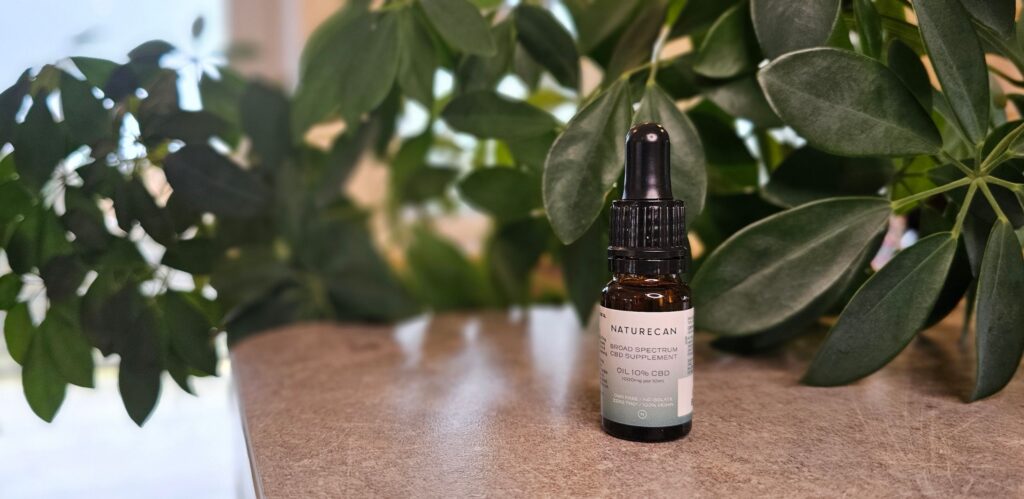 En flaska Naturecan CBD olja 10% står på en grå bänkskiva med gröna blad i bakgrunden. Flaskan är tillverkad i brunt glas, har en svart pipett och vit-grön etikett med grön text.