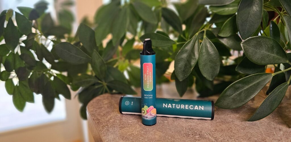 En CBD vape från Naturecan står placerad på en grå bänkskiva. Bakom den ligger dess förpackning och i bakgrunden syns gröna växter. 