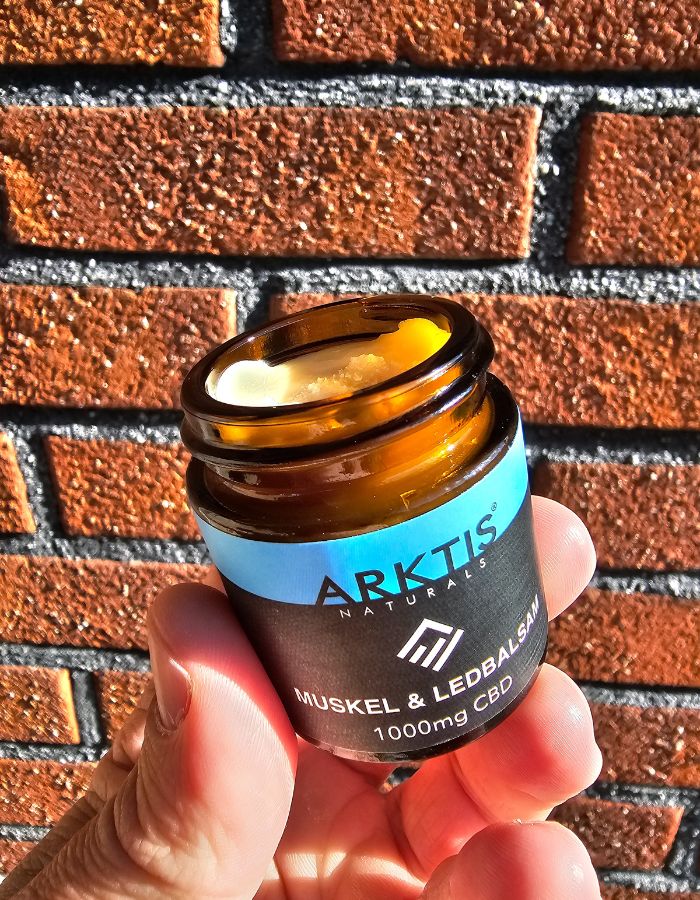 Öppnad burk med CBD balsam från arktis naturals hålls upp framför en tegelvägg. 