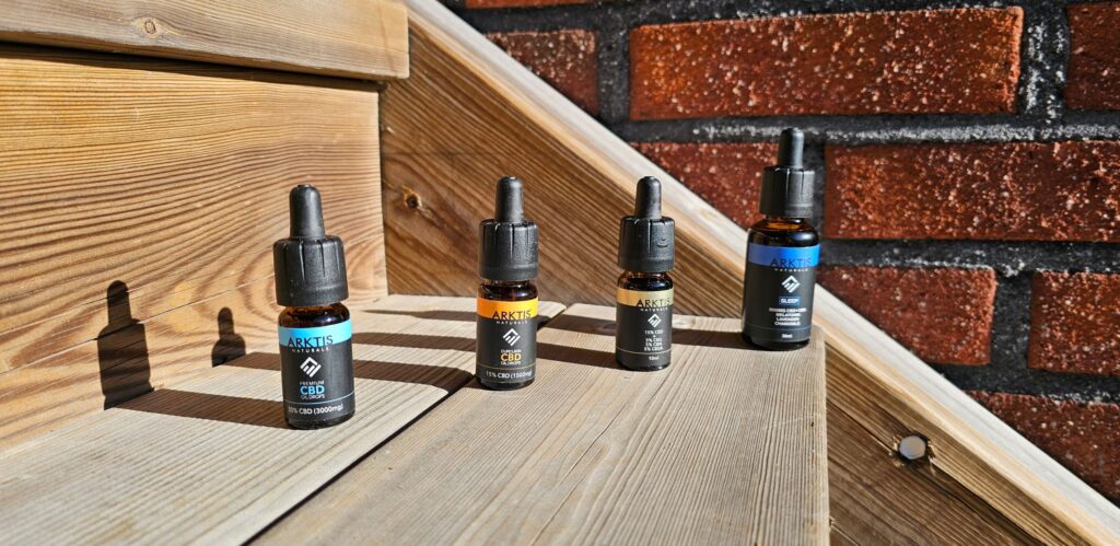 4 flaskor med olika typer av CBD olja från Arktis Naturals står placerade i en trätrappa med en tegelvägg i bakgrunden.