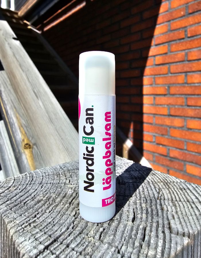 Ett vitt CBD läppbalsam från Nordic Med Can placerat stående på en trästolpe. I bakgrunden syns en tegelvägg och en trappa.